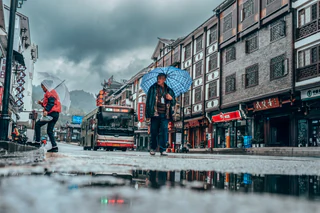 Two men carrying umbrellas cross a rain-wet street as a bus passes behind them — UNSTAGED · LOG /013 · MOMENT /006 · ARCHIVE ID L013-M006.