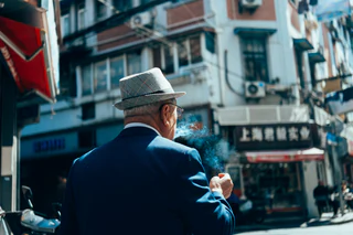 An older man in a checked hat exhales cigarette smoke on a city street — UNSTAGED · LOG /013 · MOMENT /002 · ARCHIVE ID L013-M002.