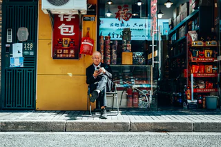 A seated man checks his phone outside a brightly coloured shop — UNSTAGED · LOG /013 · MOMENT /003 · ARCHIVE ID L013-M003.