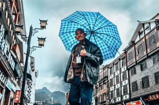 An older man walks beneath a blue checked umbrella on a mountain-town street — UNSTAGED · LOG /013 · MOMENT /007 · ARCHIVE ID L013-M007.