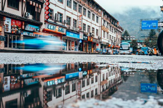 Motion-blurred vehicles pass shops reflected in a rain puddle, with misty mountains beyond — UNSTAGED · LOG /013 · MOMENT /005 · ARCHIVE ID L013-M005.